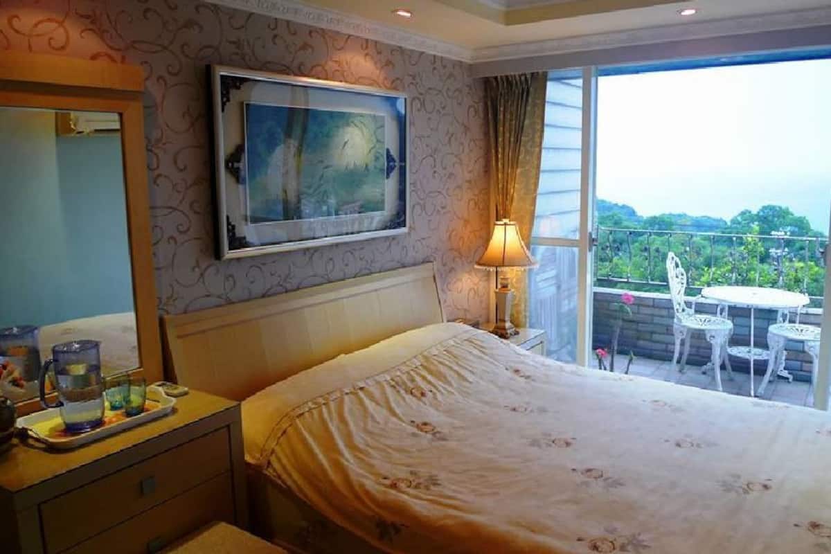 Une chambre d’hôtel avec un lit, une table de chevet avec une lampe, un tableau au mur et un balcon avec une table et des chaises.