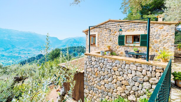 Une maison en pierre avec un mur de pierre et une porte en bois, un petit patio avec une table et des chaises, et une vue sur les montagnes.