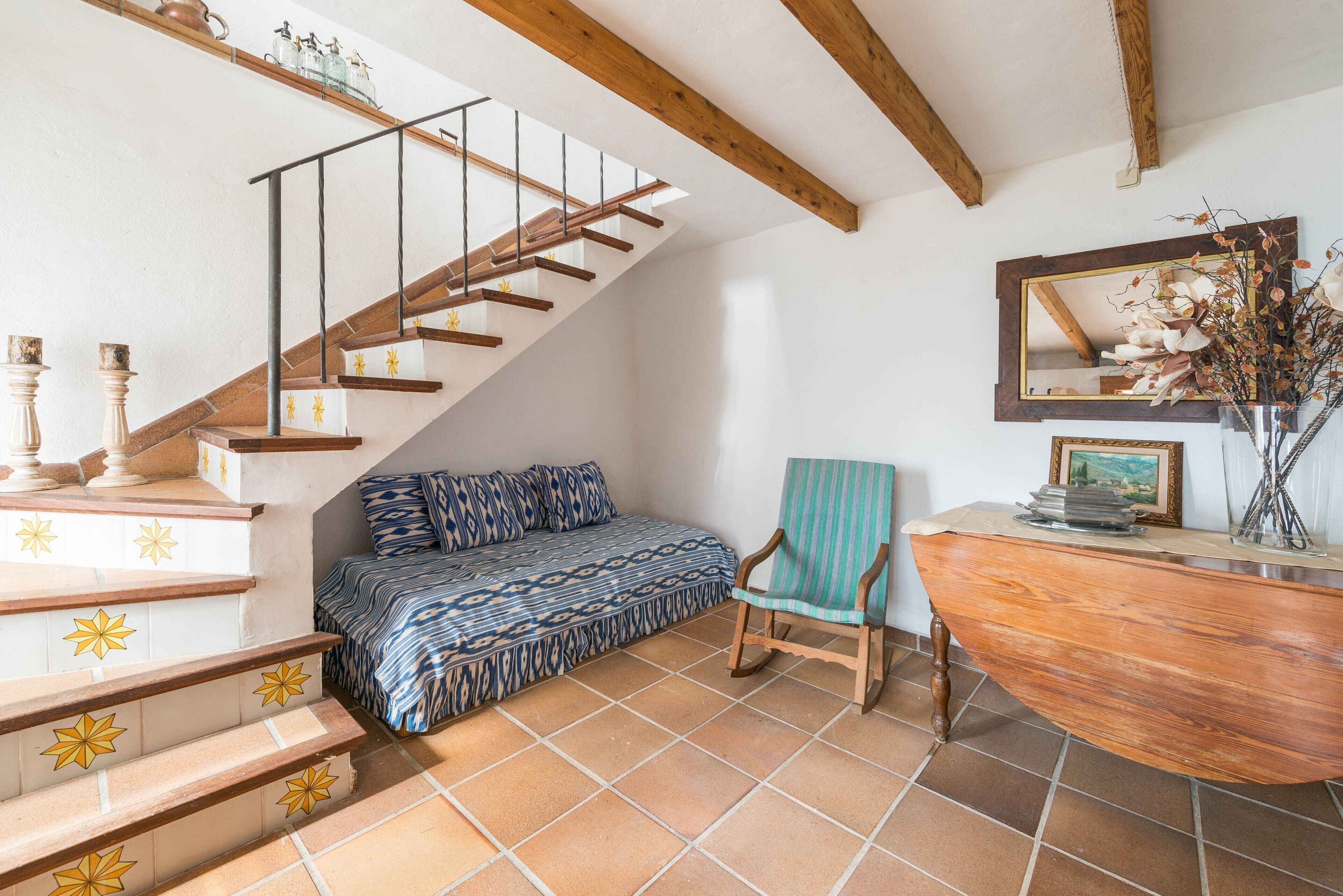 A bedroom with a wooden staircase, a bed with patterned bedding, a chair, and a desk with a mirror and vase.