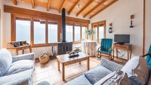 Un salon chaleureux avec des poutres apparentes, de grandes fenêtres, une cheminée, un téléviseur et des fauteuils confortables.