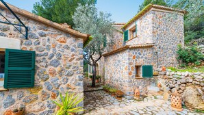 Un bâtiment aux murs de pierre, avec un étroit sentier pavé, des volets verts et des plantes en pot.