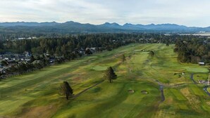 An aerial view of a golf course with a forested area, a few scattered houses, and mountains in the background.