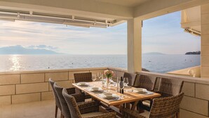 A dining area with a view of the sea, featuring a wooden table set for four, wicker chairs, and a large window overlooking the ocean.