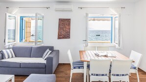 A room with a grey sofa, a wooden dining table, and wicker chairs. There are open windows with views of the sea.