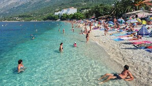 A crowded beach with clear turquoise water, people swimming and sunbathing, and a mountainous backdrop.