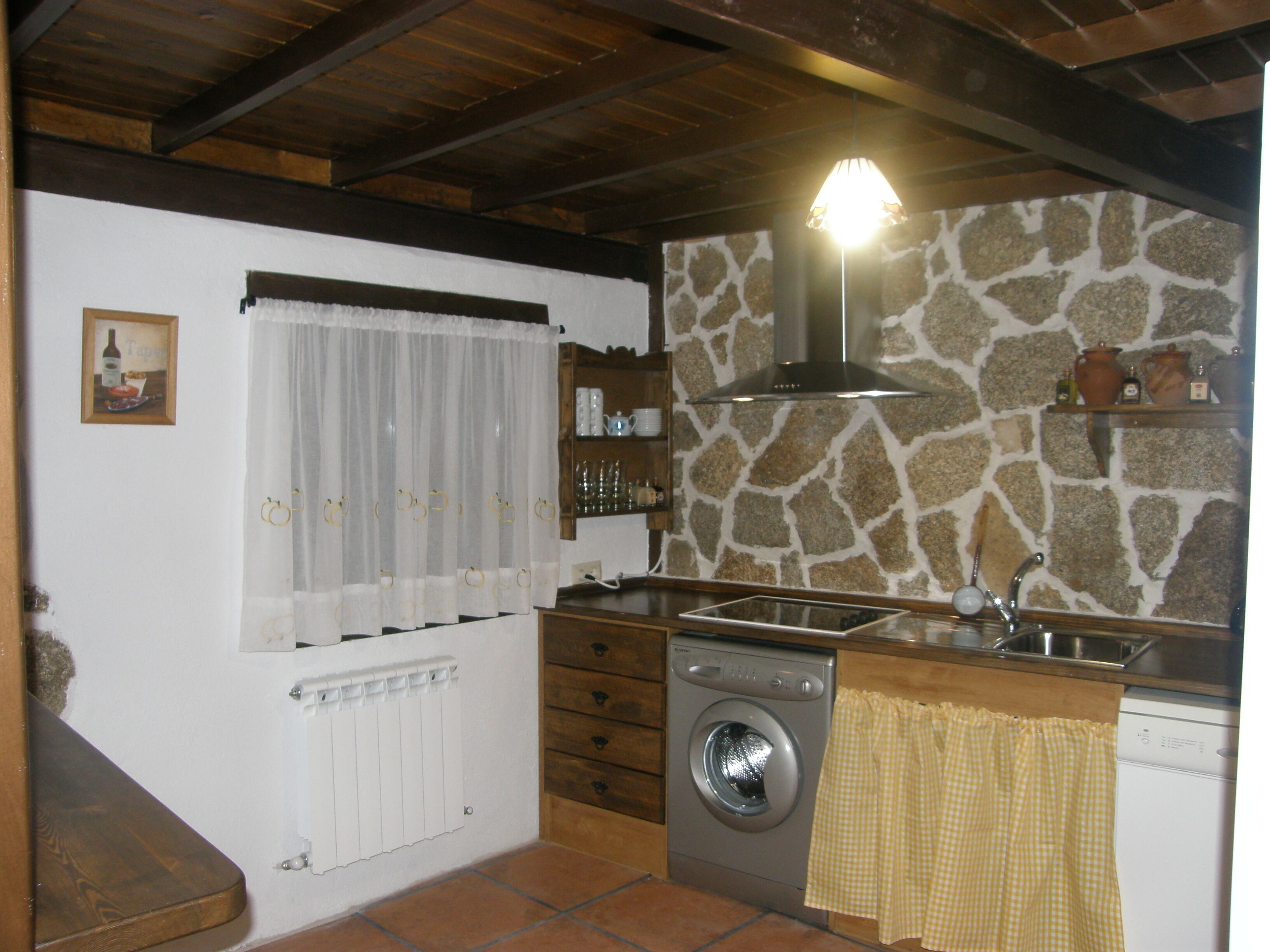 A kitchen with a stone wall, a washing machine, a sink, and a window with curtains.