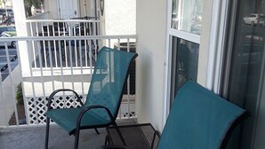 Ein Balkon mit zwei sesseln in Türkis und einem Tisch, eine weiße Tür und Blick auf geparkte Autos.