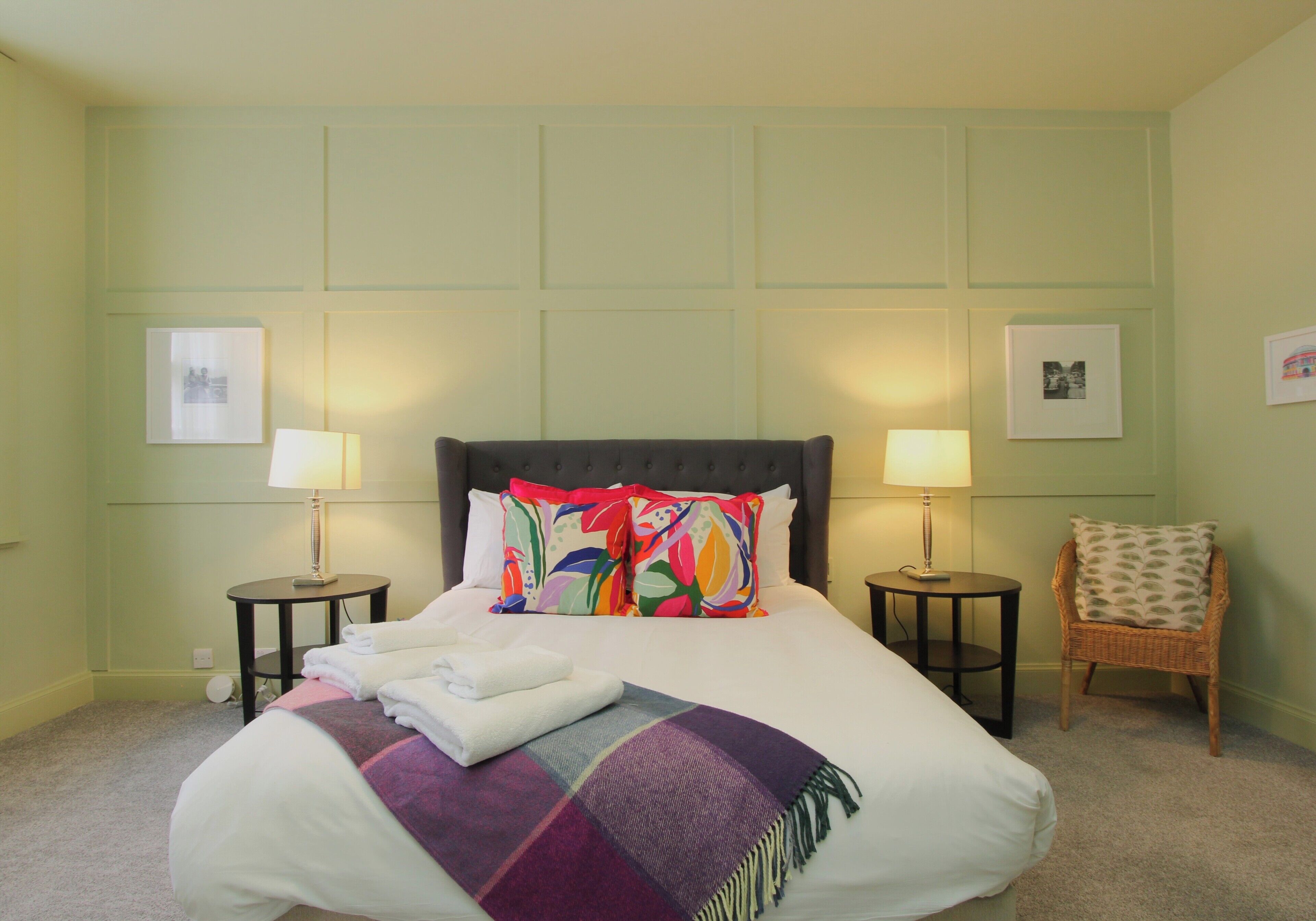 A neatly made bed with a colorful blanket and pillows, two bedside tables with lamps, and a wicker chair in a room with light green walls.