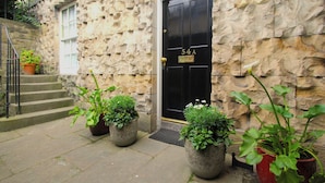 Un bâtiment en pierre avec une porte noire portant l’inscription « 54A » et un escalier menant à une porte dotée d’une fenêtre. Des plantes en pot sont disposées au sol.