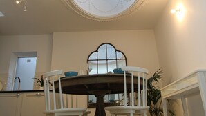 Un coin repas avec une table ronde, deux chaises blanches, une grande fenêtre ornée d’un cadre décoratif et un plafond doté d’une lucarne.