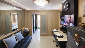 A modern hotel room with a sofa, a dining table with plates and glasses, a TV displaying a movie, and a clock on the wall. - 1 BR Fully Furnished Condo Across MOA with Pool and Parking - Shore Bldg B, 1244 (Pasay City)