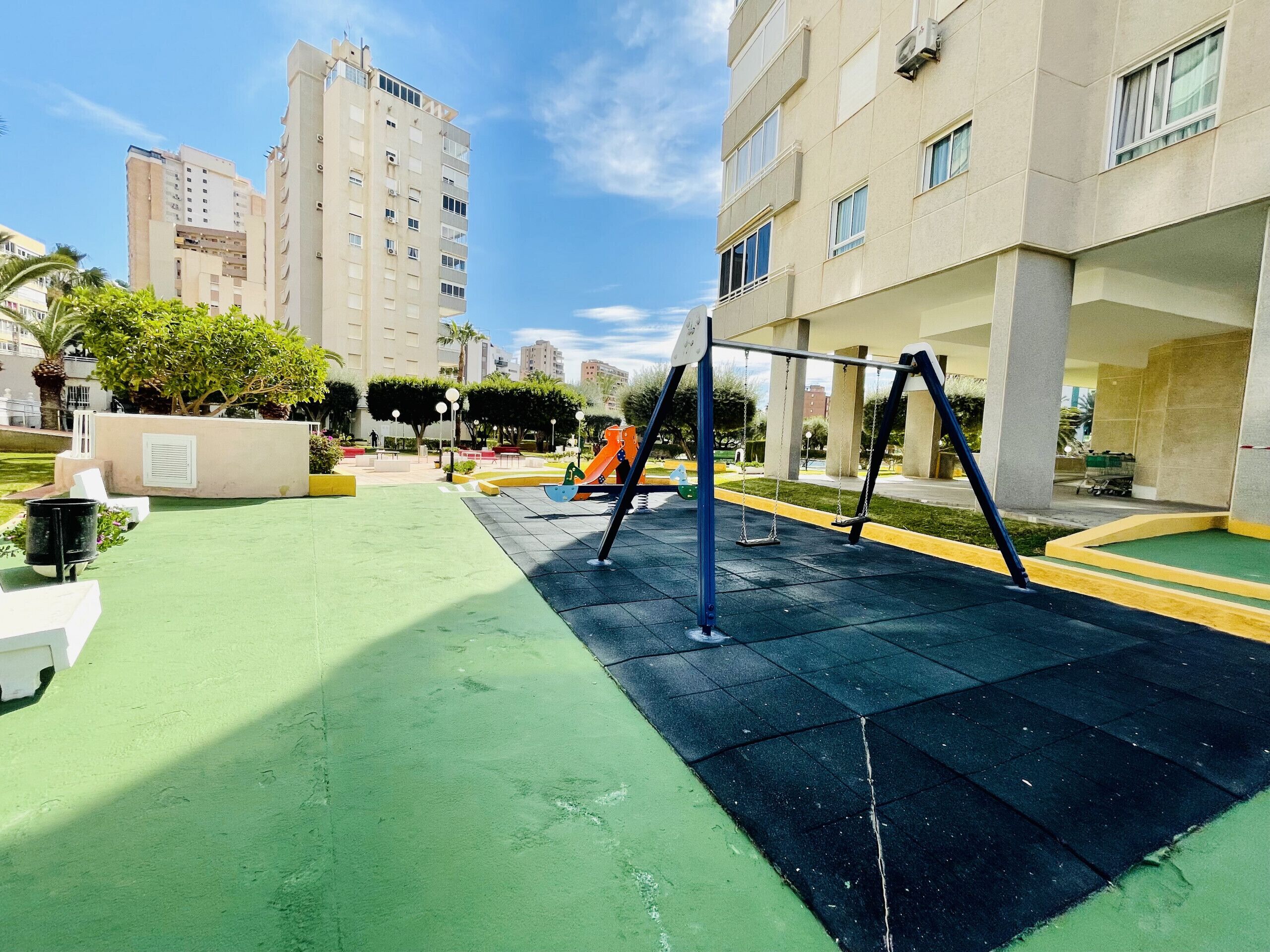 A playground with swings and a slide, surrounded by buildings and greenery.