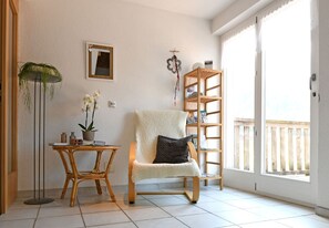 Ein Zimmer mit einem Sessel, einem Tisch mit einer Pflanze, einem Bücherregal und einem Fenster mit Blick.
