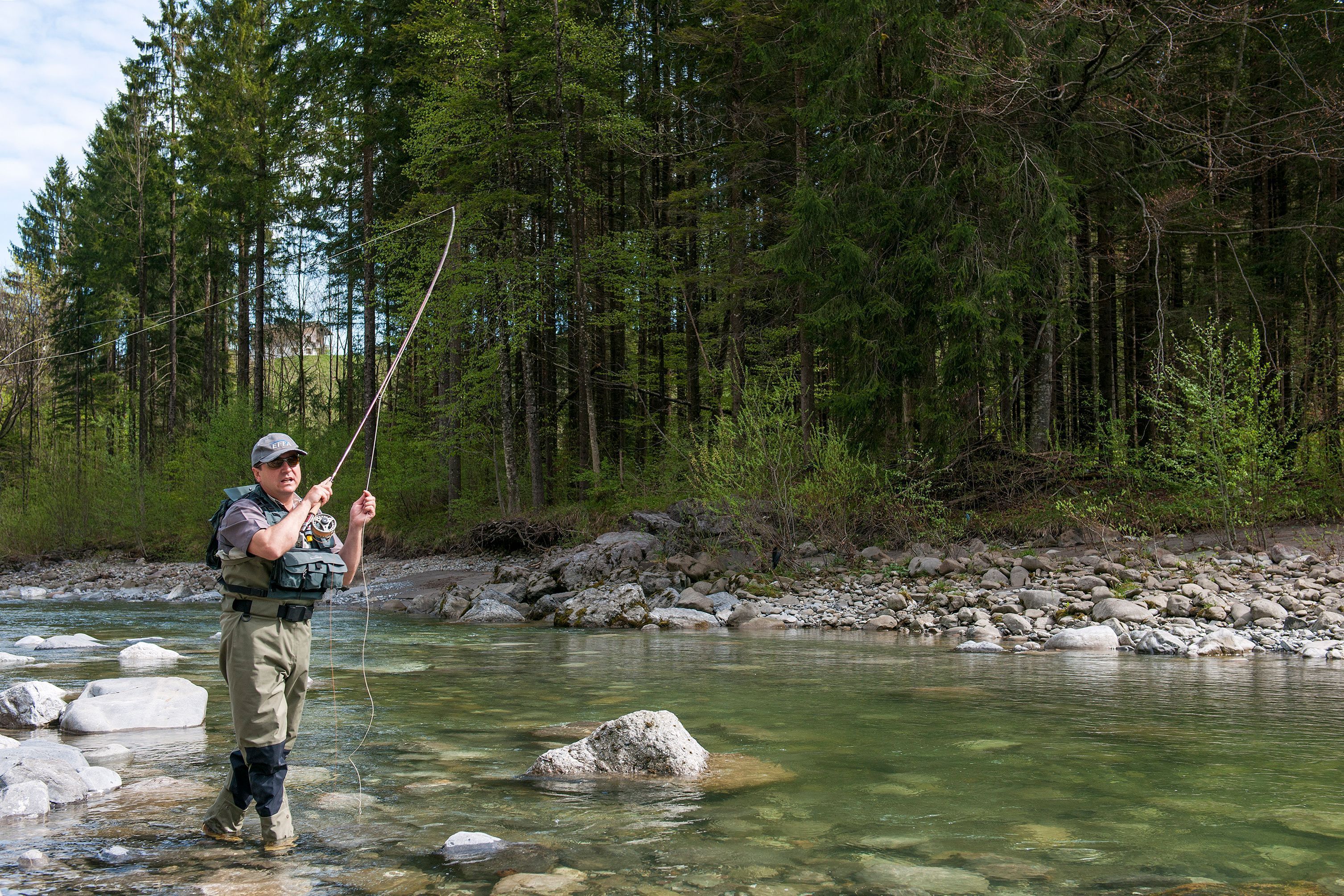 A person standing in a clear river with rocks, surrounded by dense forest.
