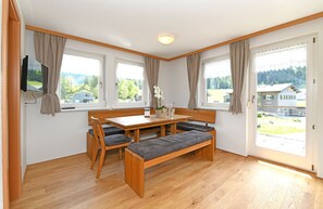 A room with a wooden table, benches, a TV, and large windows with curtains.