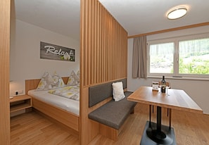 Een kleine kamer met een bed, een bankje, een tafel en een raam met uitzicht.