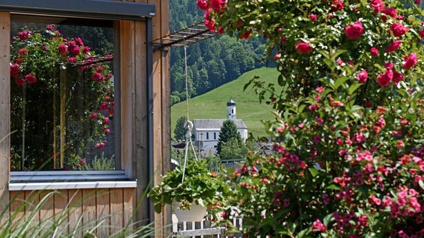 A wooden building with a window overlooking a green landscape and a church.