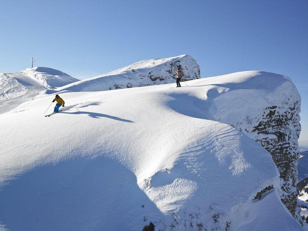 Eine verschneite Berglandschaft mit einem Kreuz auf einem Hügel und einer Person beim Skifahren.