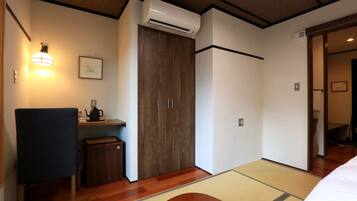 A hotel room with a wooden floor, a bed, a desk with a chair, a small cabinet, a wardrobe, and an air conditioning unit.