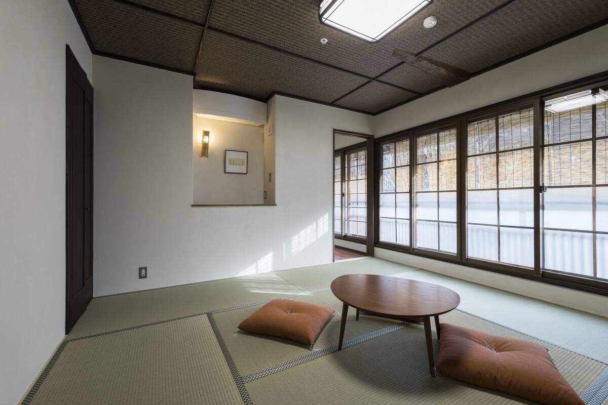 A traditional Japanese room with tatami flooring, a low table, cushions, and large windows with a view.