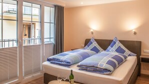 A bedroom with a bed, two bedside tables, a small round table with wine glasses and a bottle, and a balcony with blinds. - Das Gästehaus in Valwig (Valwig)