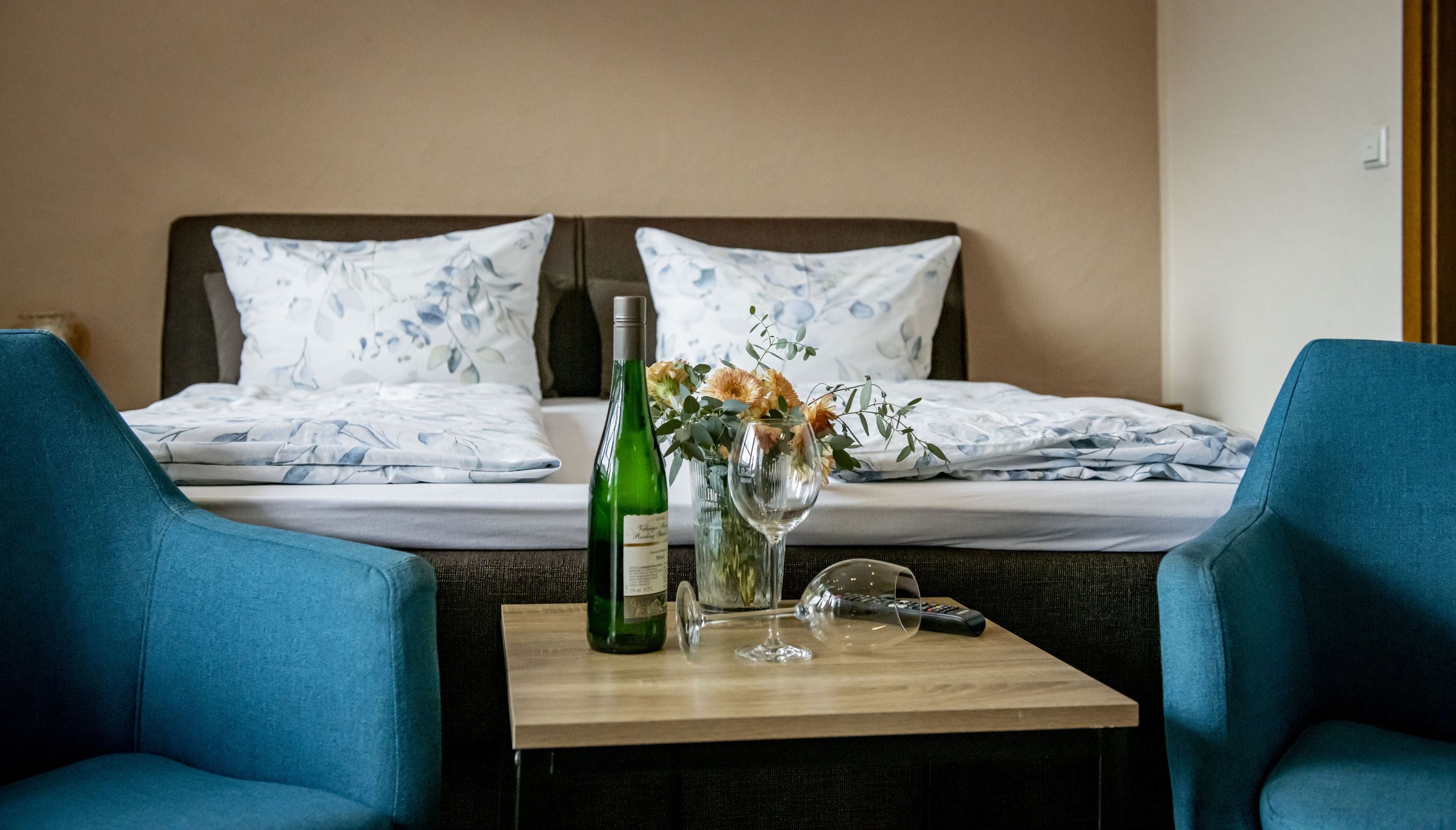 A hotel room with two beds, a small table with a bottle and a vase of flowers, and two blue armchairs.
