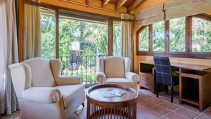 Une pièce avec deux fauteuils beiges, un bureau en bois et une table basse ronde. De grandes fenêtres offrent une vue sur la verdure environnante.