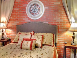 A bedroom with a brick wall, a round clock, a bed with floral bedding, and two bedside tables with lamps.
