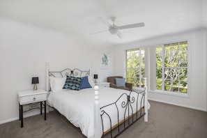 Une chambre avec un lit blanc, un ventilateur de plafond, une fenêtre donnant sur des arbres et une table de chevet avec une lampe.