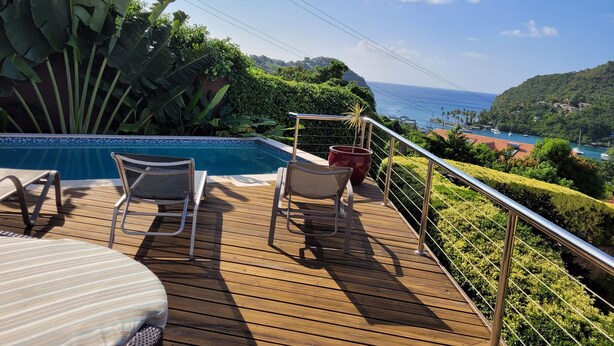 Una terraza de madera con piscina, dos sillones de descanso y vistas al océano y colinas.