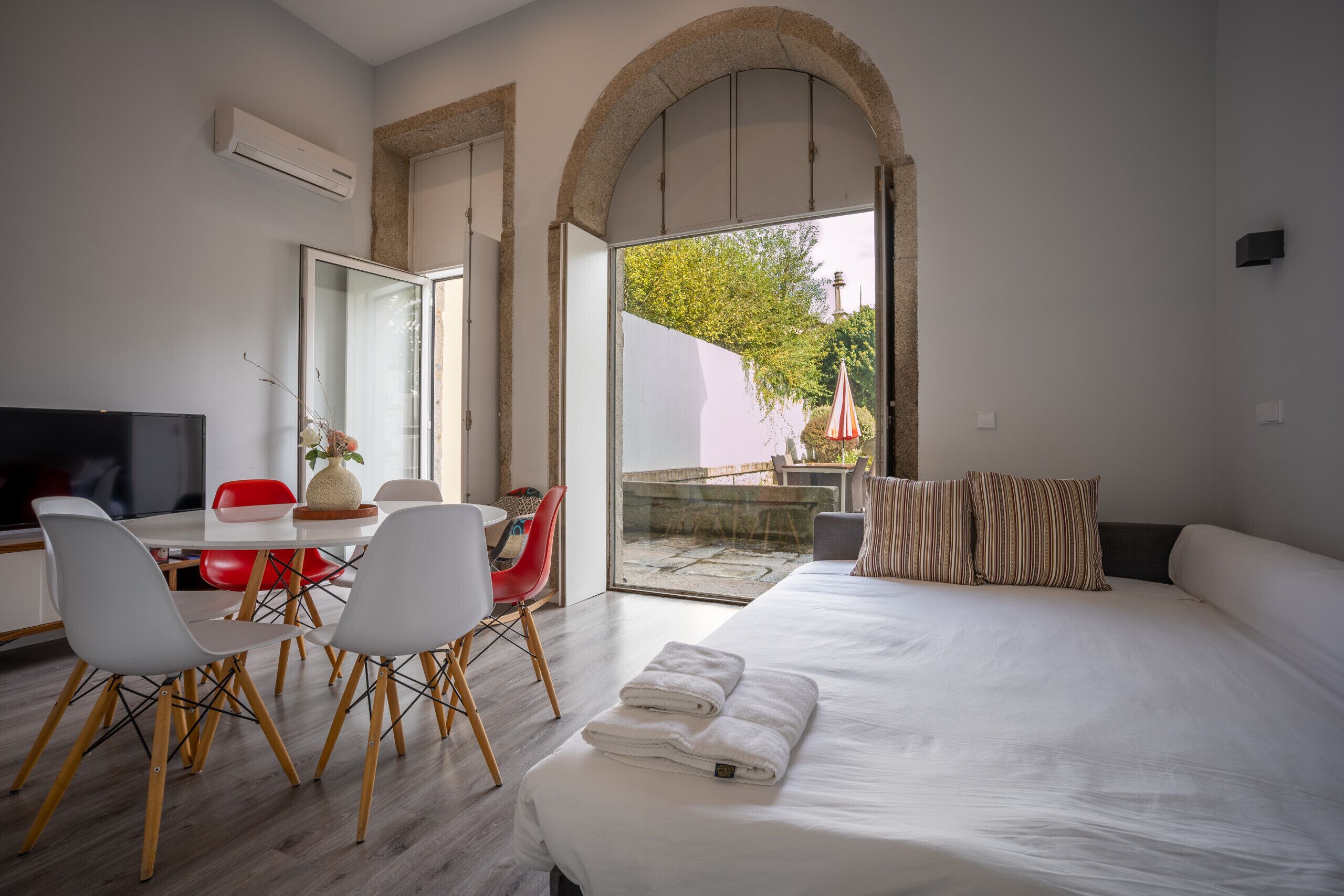 A modern hotel room with a large bed, a dining area with a table and chairs, a TV, and a view through an open archway to an outdoor area.