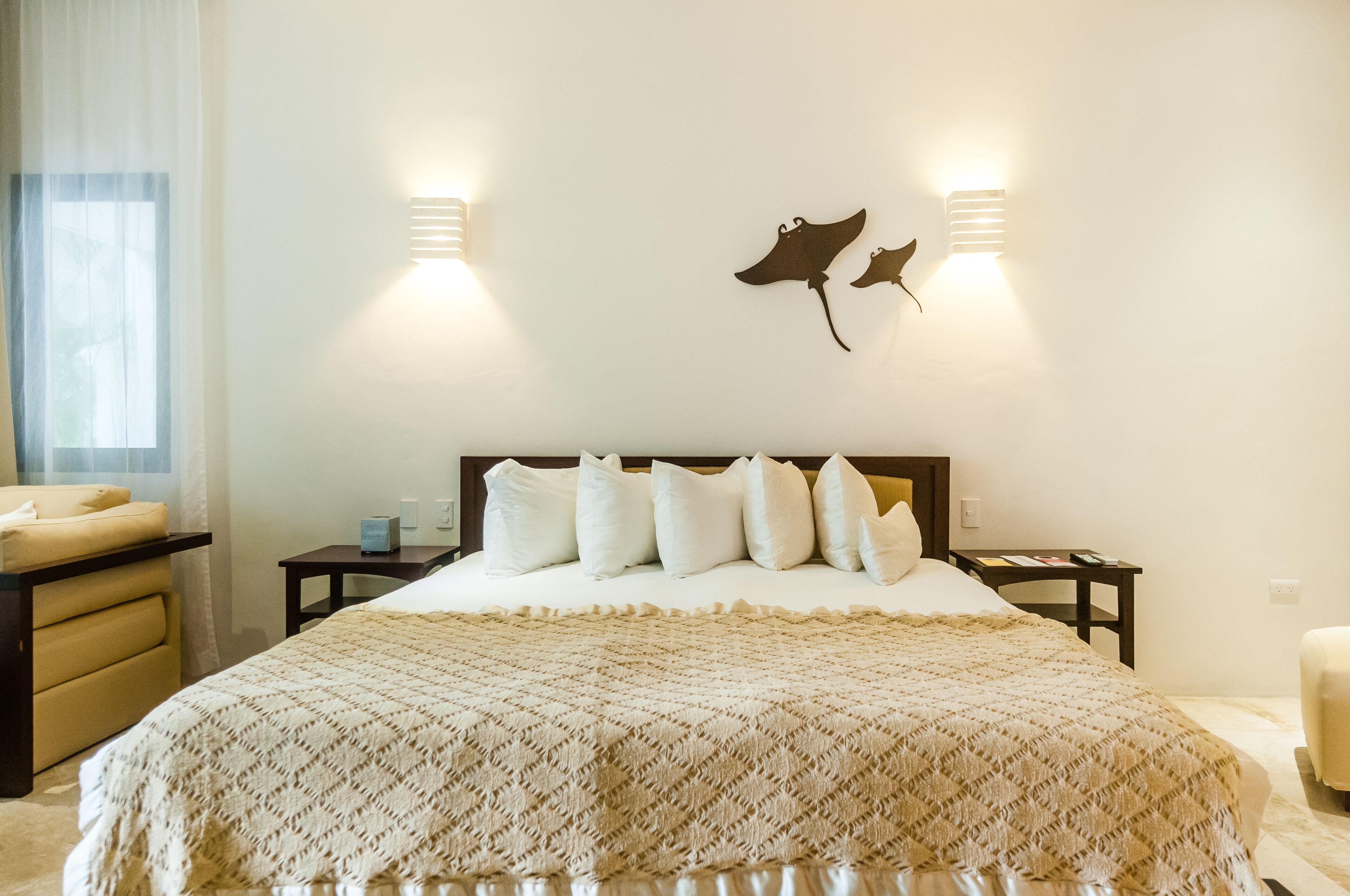 Een keurig opgemaakt bed met een bedsprei met patroon, een nachtkastje met een lamp, en een wandlamp met een motief van een mantaray.