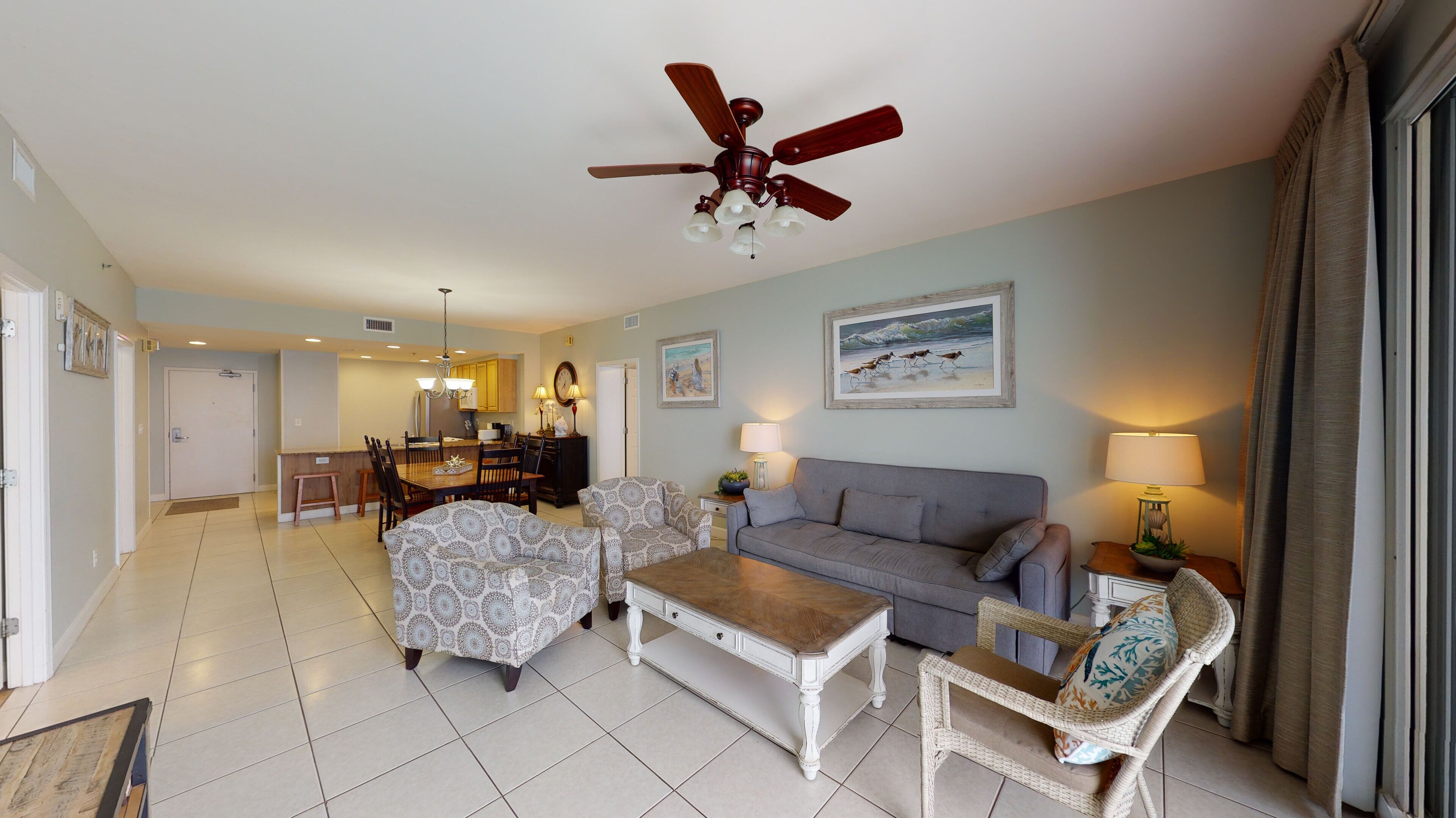A spacious living room with a grey sofa, patterned armchairs, a wooden coffee table, and a ceiling fan.