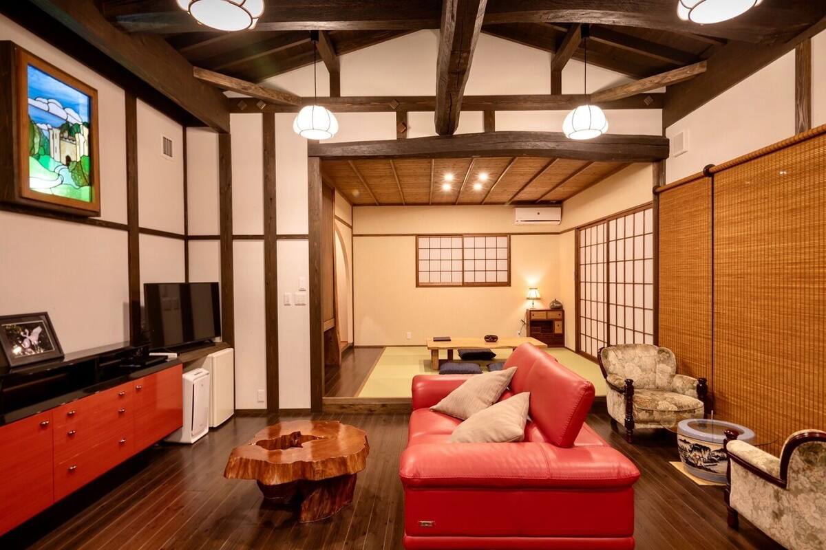 Ein traditioneller japanischer Wohnraum mit Tatami-Böden, einem roten Sofa, einem Holztisch und einem Fernseher auf einem Schrank.