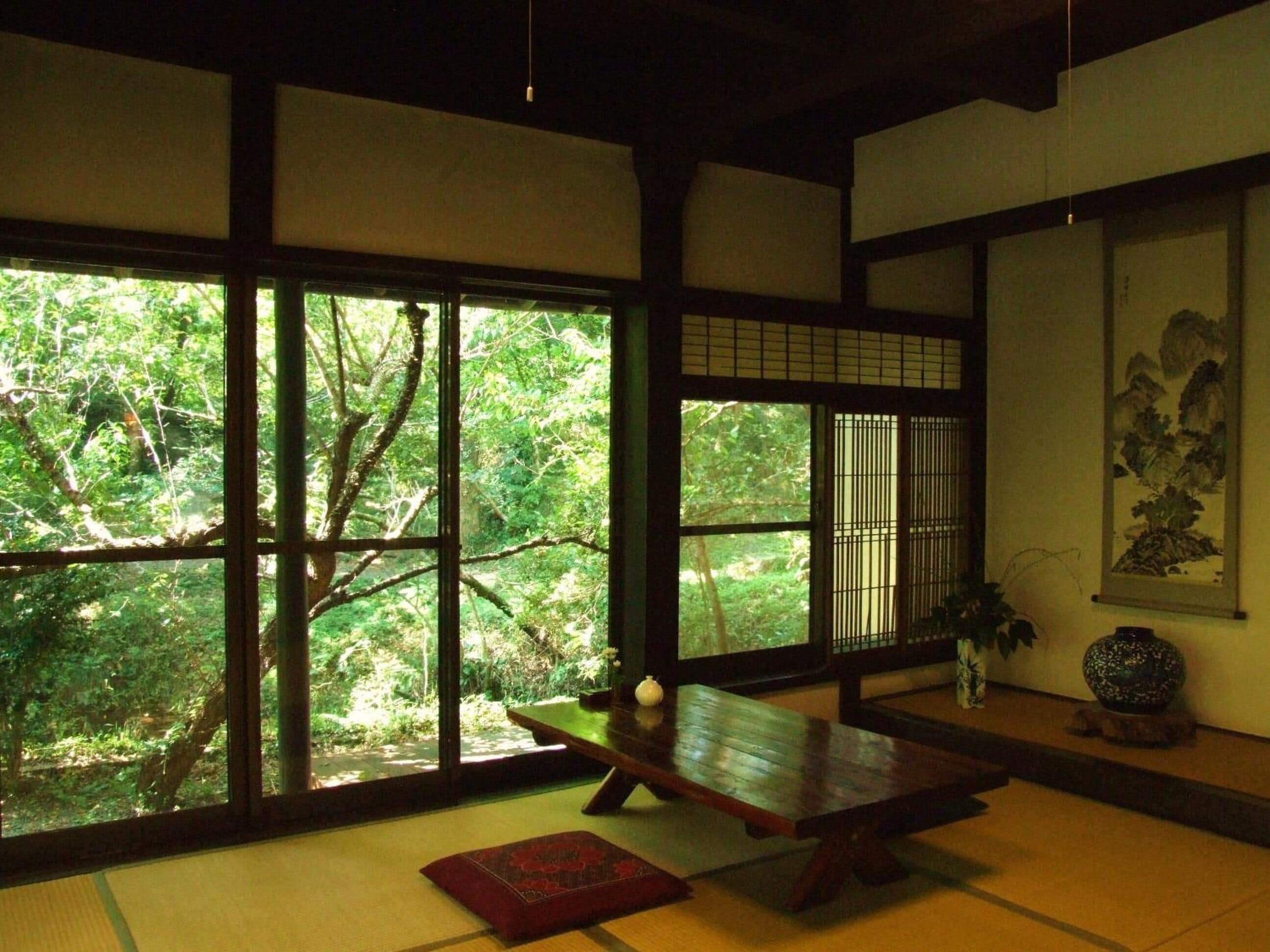 A traditional Japanese room with tatami flooring, a low wooden table, and large windows offering a view of a lush garden.
