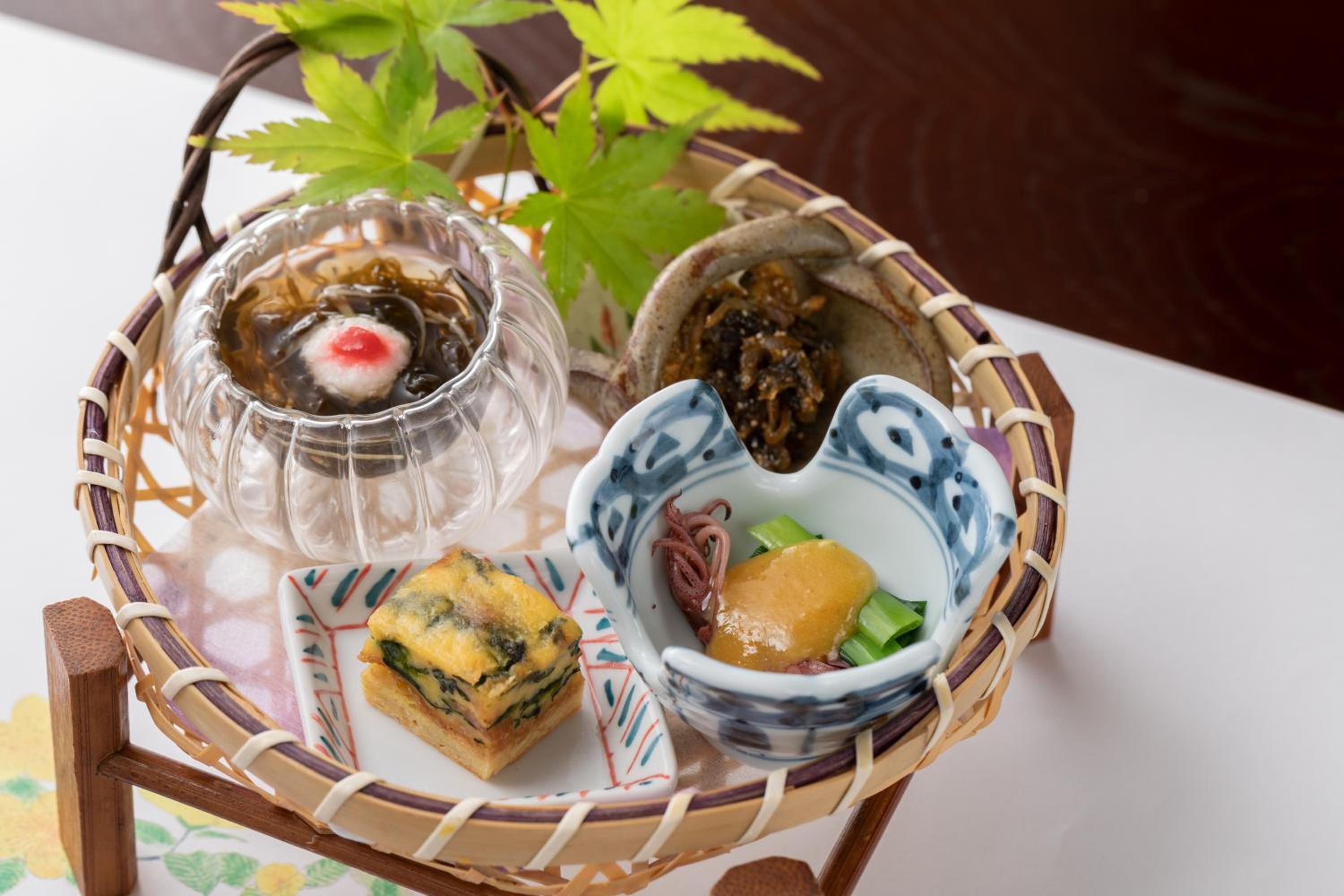 A traditional Japanese meal set on a woven tray, including a clear glass bowl with a red center, a ceramic bowl with a blue pattern, and a small plate with a green and yellow dish.