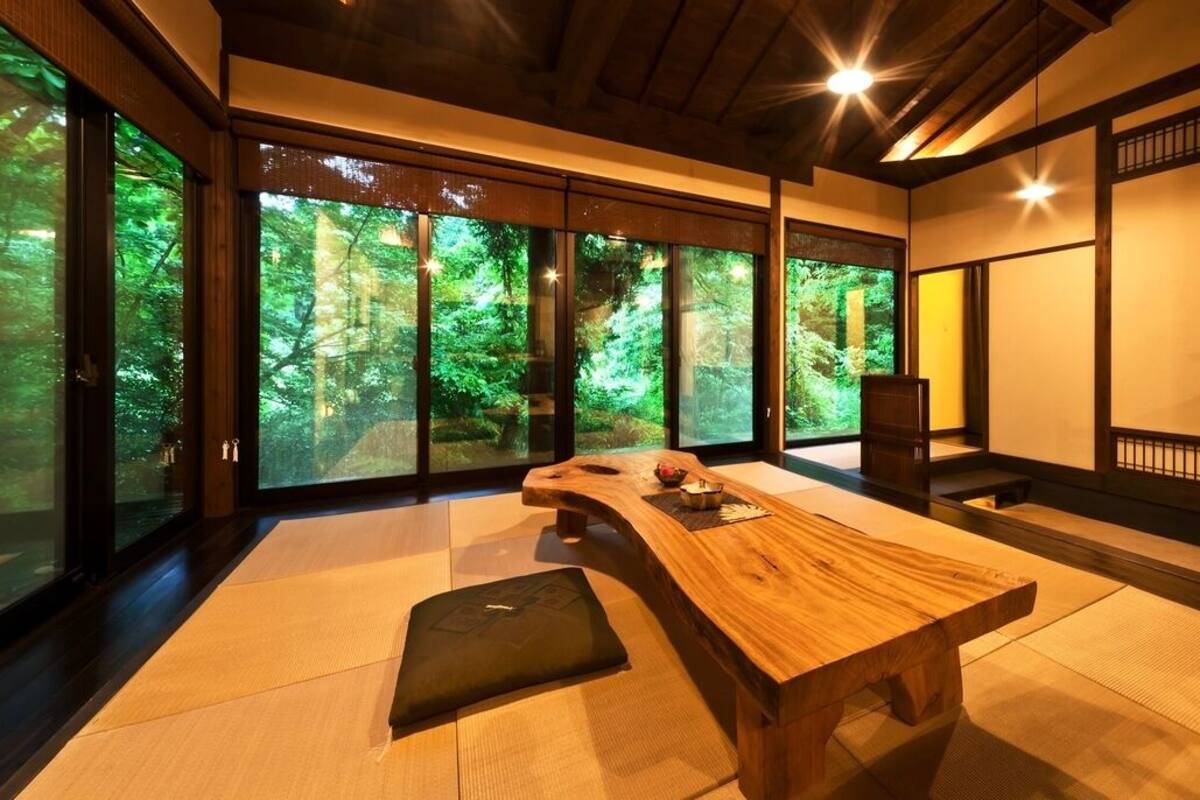 Ein traditioneller japanischer Raum mit Tatami-Böden, einem niedrigen Holztisch, Schiebetüren mit Blick auf einen Wald und einem dunklen Holzstuhl.