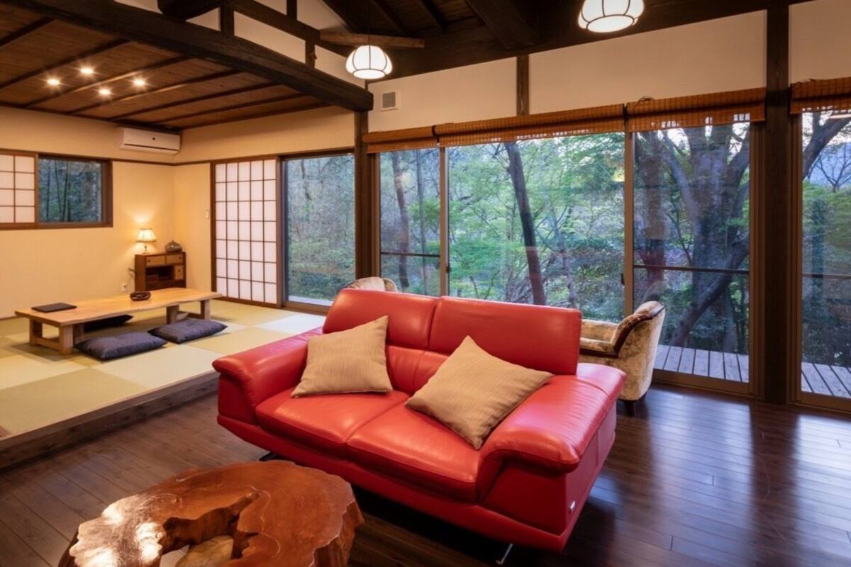 Ein Zimmer mit einem roten Ledersofa, Holzdielenboden, großen Fenstern mit Blick auf Bäume und einer traditionellen japanischen Lampe.