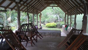 A covered outdoor seating area with wooden chairs and tables, surrounded by lush greenery and a stone bench.