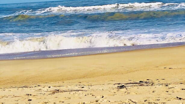 Ein Strand mit Wellen, einem sandigen Ufer und einem Segelboot in der Ferne.