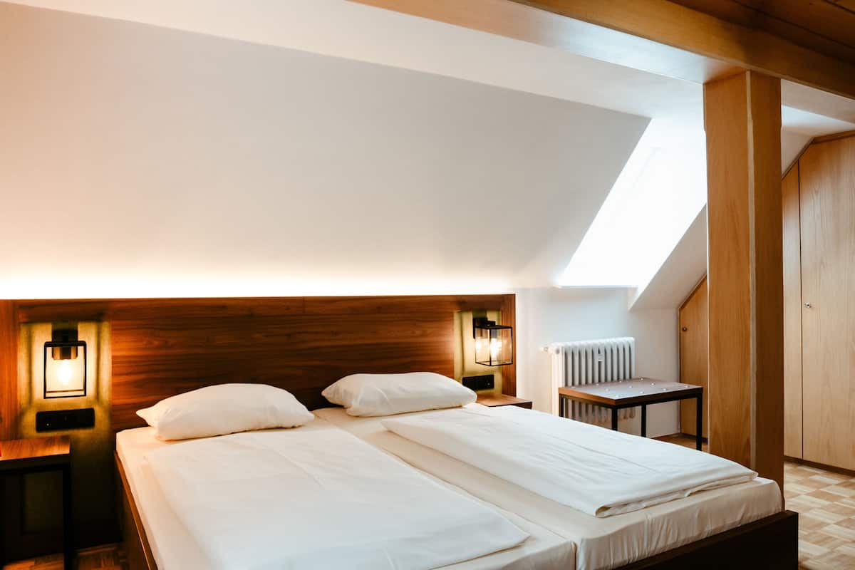 Une chambre avec deux lits, une tête de lit en bois, des lampes de chevet, un radiateur et une lucarne.