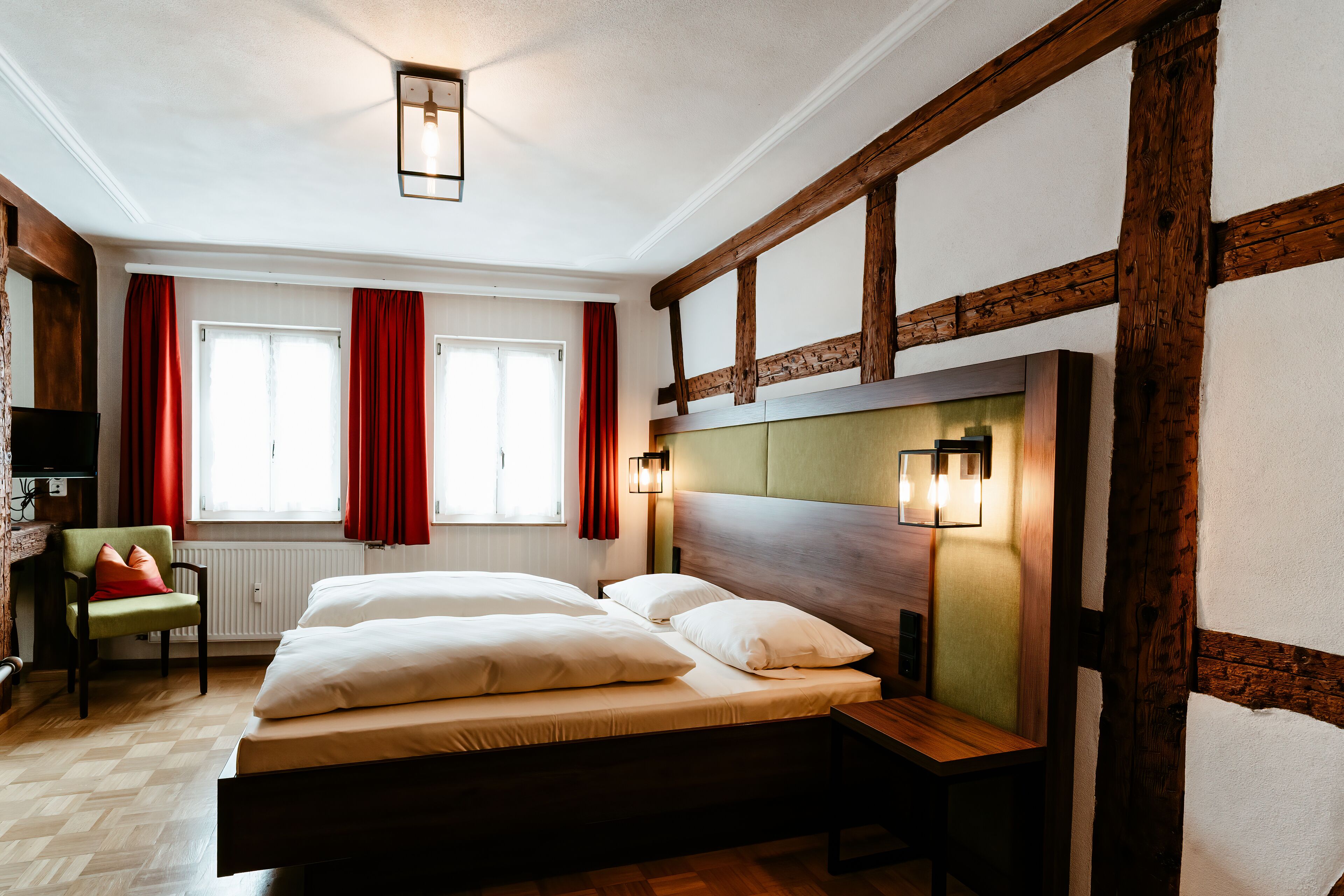 A room with a large bed, wooden beams, red curtains, and a green chair.
