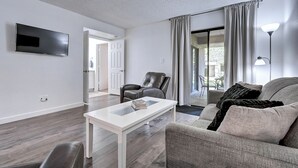 Ein modernes Wohnzimmer mit einem Flachbildfernseher, einem weißen Couchtisch, einem Ledersessel, einem grauen Sofa und einem großen Fenster mit Vorhängen.