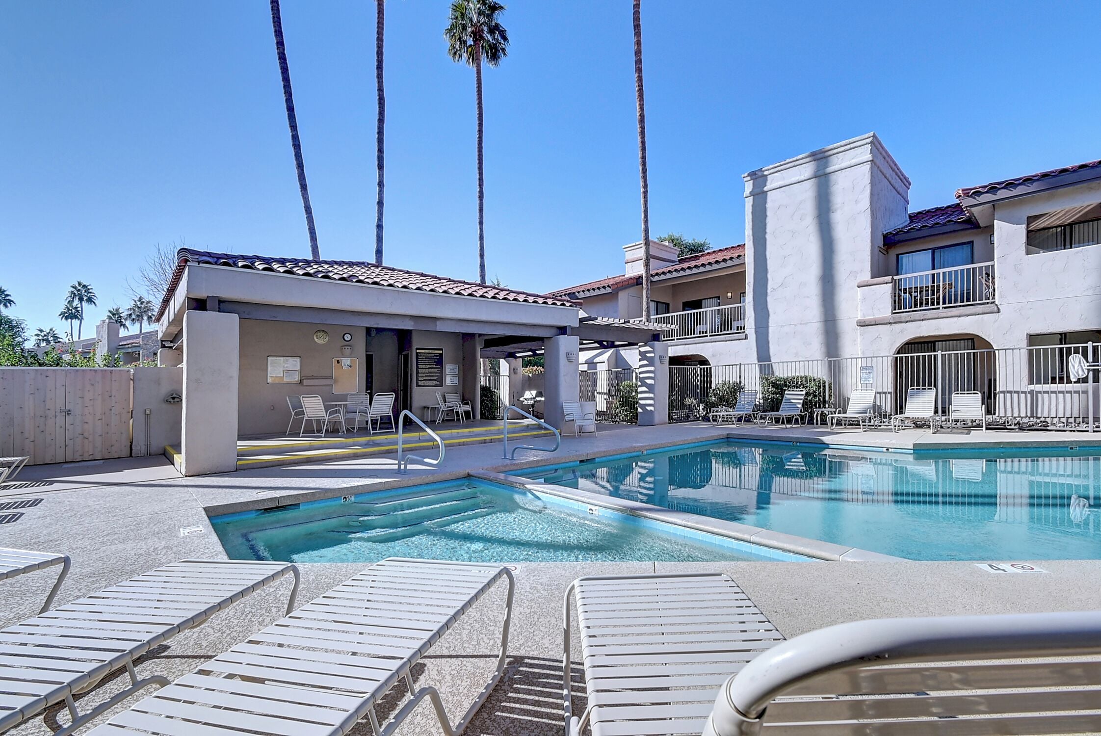 A pool area with lounge chairs, a small building with a covered seating area, and tall palm trees.