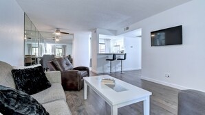 Ein modernes Wohnzimmer mit Sofa, Sessel und Couchtisch. Ein Fernseher ist an der Wand montiert, und im Hintergrund ist eine Küche zu sehen.