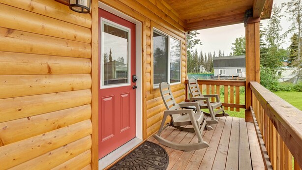 Eine Holzhütte mit roter Tür, einer Veranda mit Schaukelstühlen und Blick auf eine grüne Landschaft.