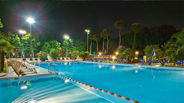 A swimming pool with lane dividers, surrounded by lounge chairs and umbrellas, under night lighting. - Hotel Caravelle (Cesenatico)