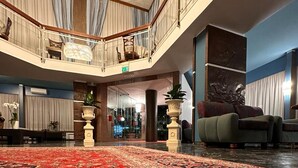 A spacious lobby with a large red patterned carpet, a seating area with sofas, a chandelier, and a view of the lobby area through glass doors. - Hotel Caravelle (Cesenatico)