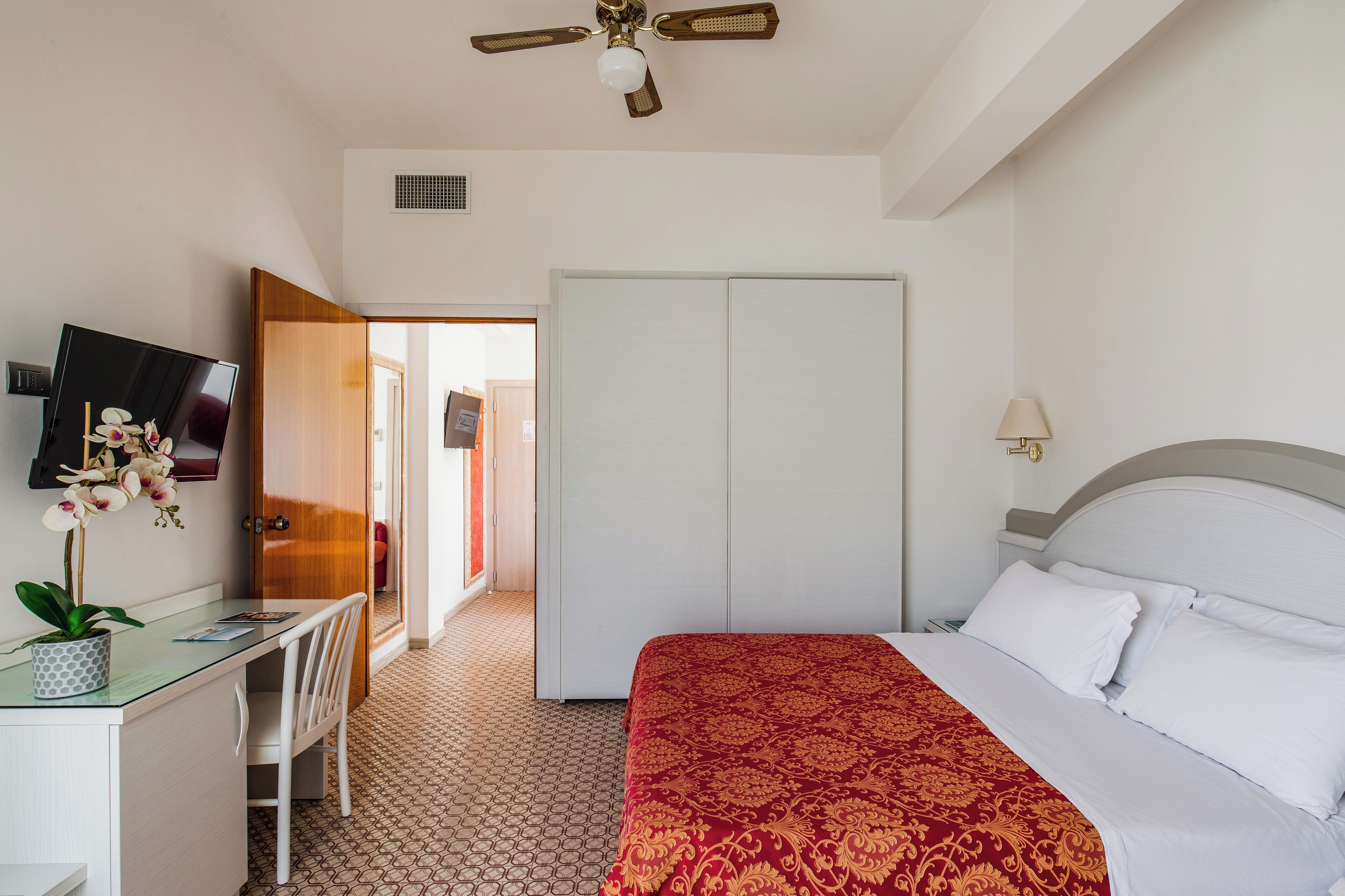 A hotel room with a bed, desk, chair, TV, wardrobe, and a ceiling fan.