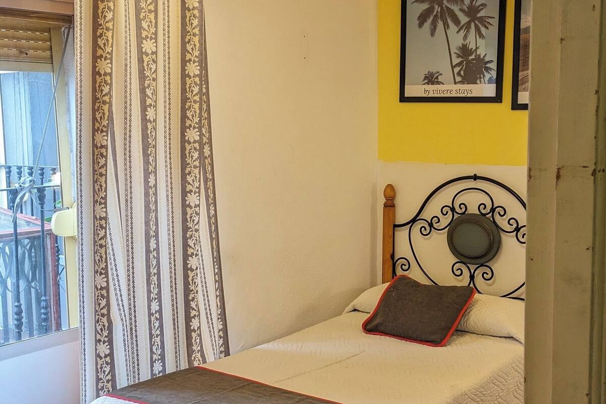 Un dormitorio con una cama, ventilador de techo, ventana con cortinas y un cuadro en la pared.